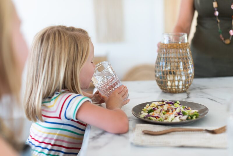 young girl drinking water at a kitchen table in front of a plate full of something